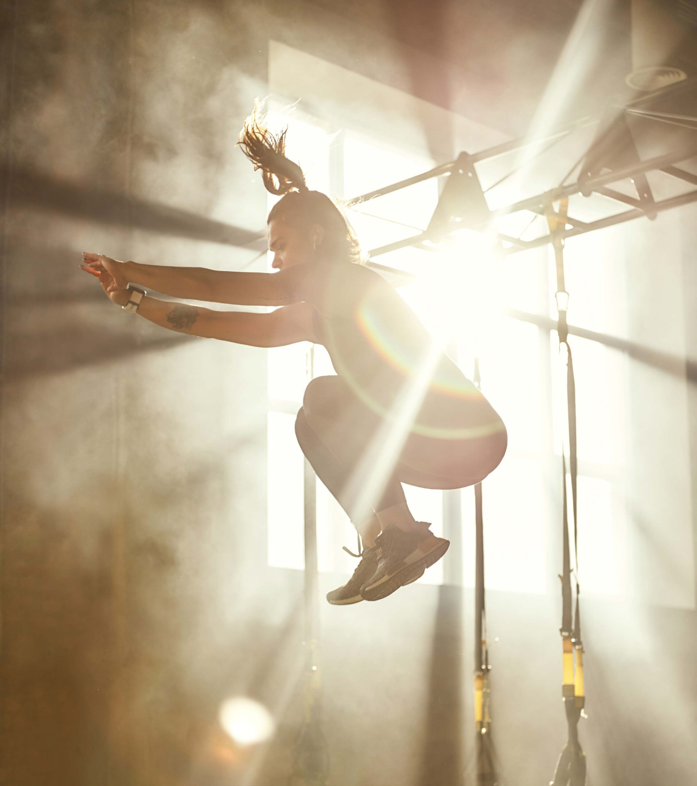A woman in a gym jumping up during a HIIT class with the sun shining through a window behind her