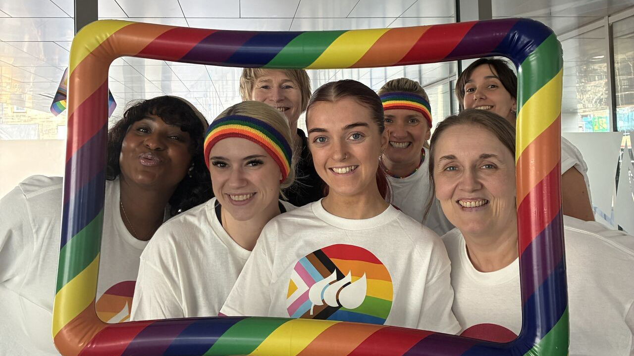 A group of people celebrating Pride, wearing rainbow motif clothing and holding an inflatable rainbow picture frame