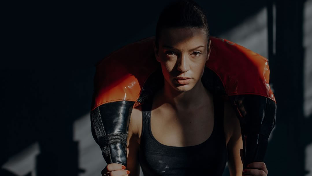 A moody looking woman in a gym with harsh lighting across her face holding a weighted bag across her sholders