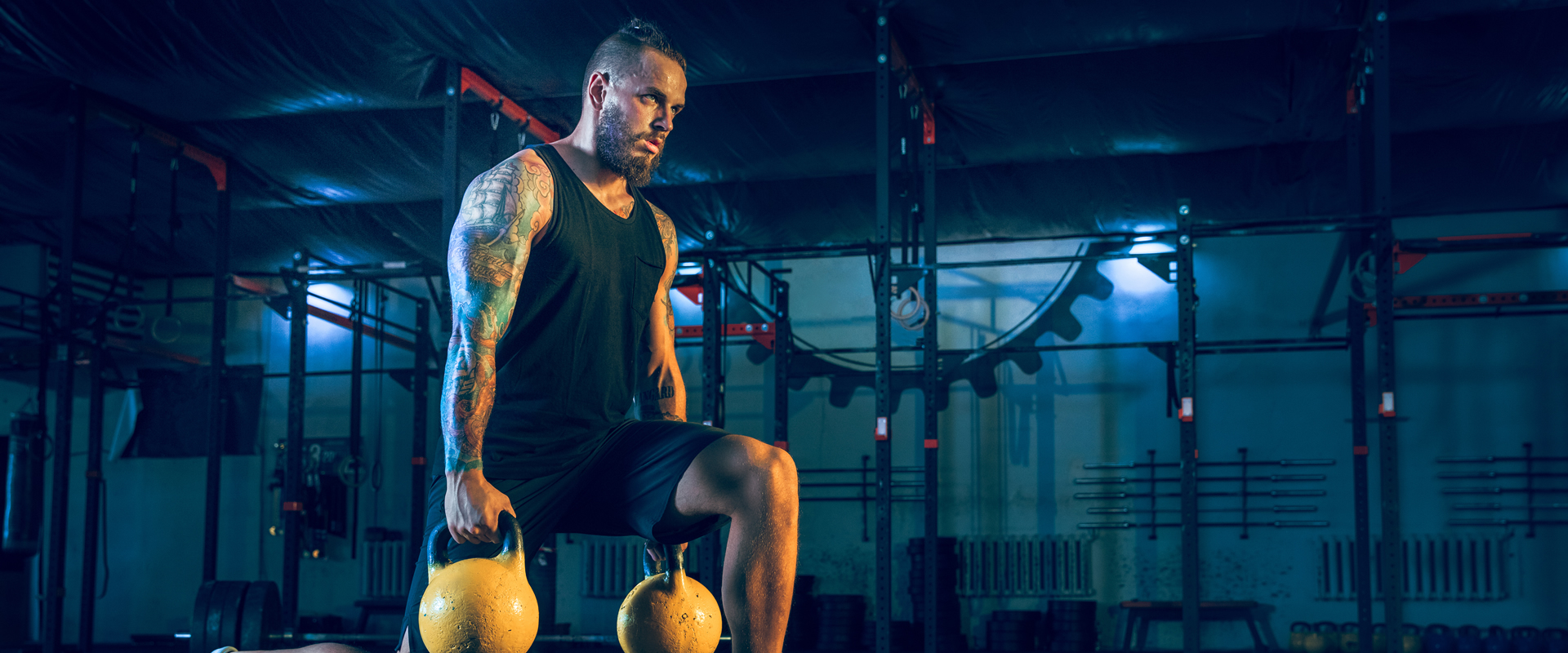Moody looking man with tattoos in a gym holding heavy kettlebells while doing lunges